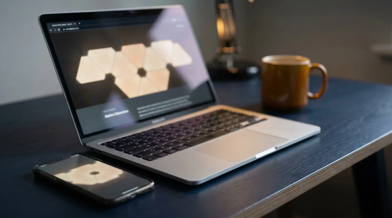 A laptop showing code on a dark desk in natural light beside a phone with an installed app icon, no people