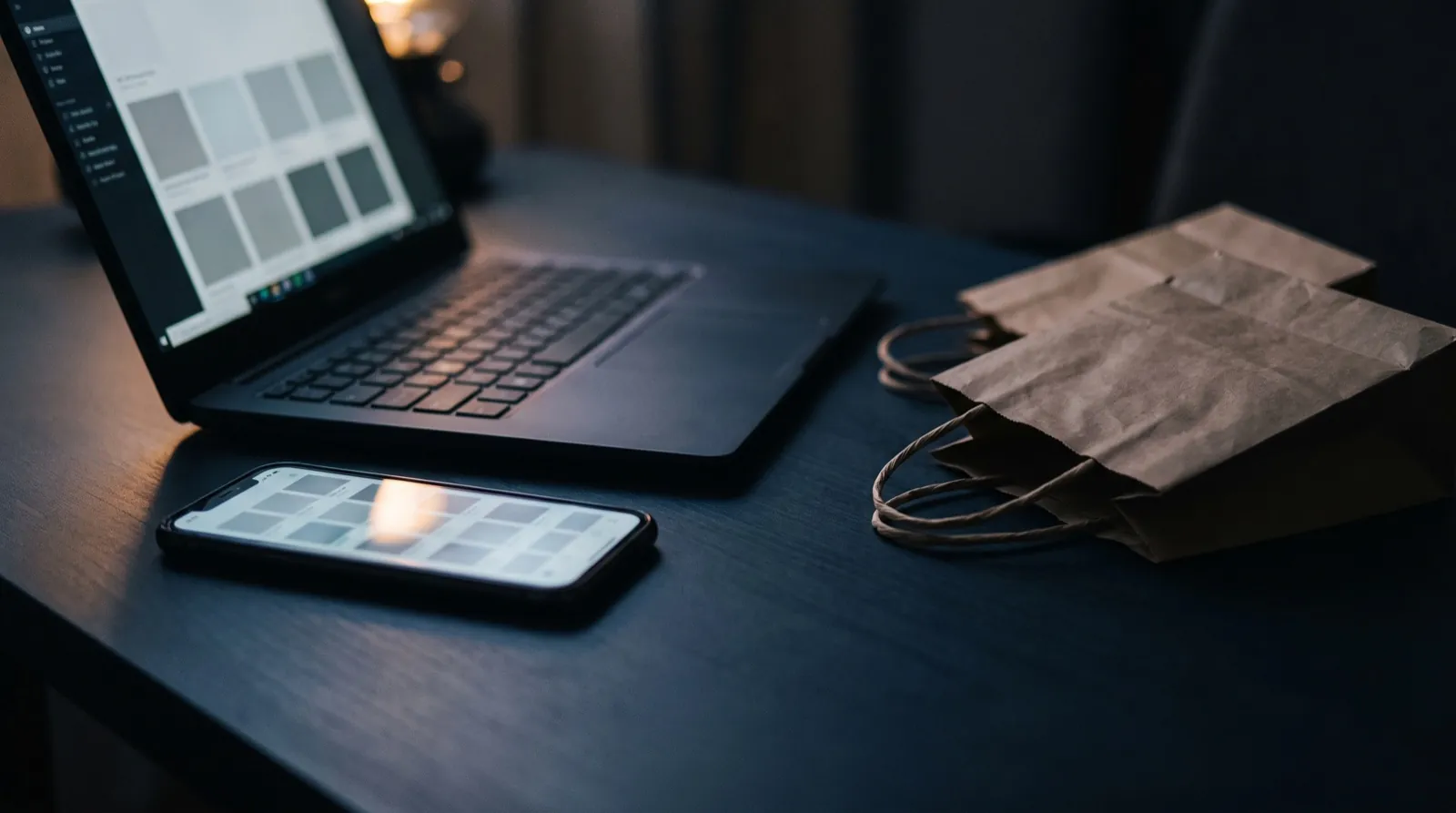A smartphone and a laptop showing a generic online store grid beside small shopping bags on a dark navy desk in low light, no people