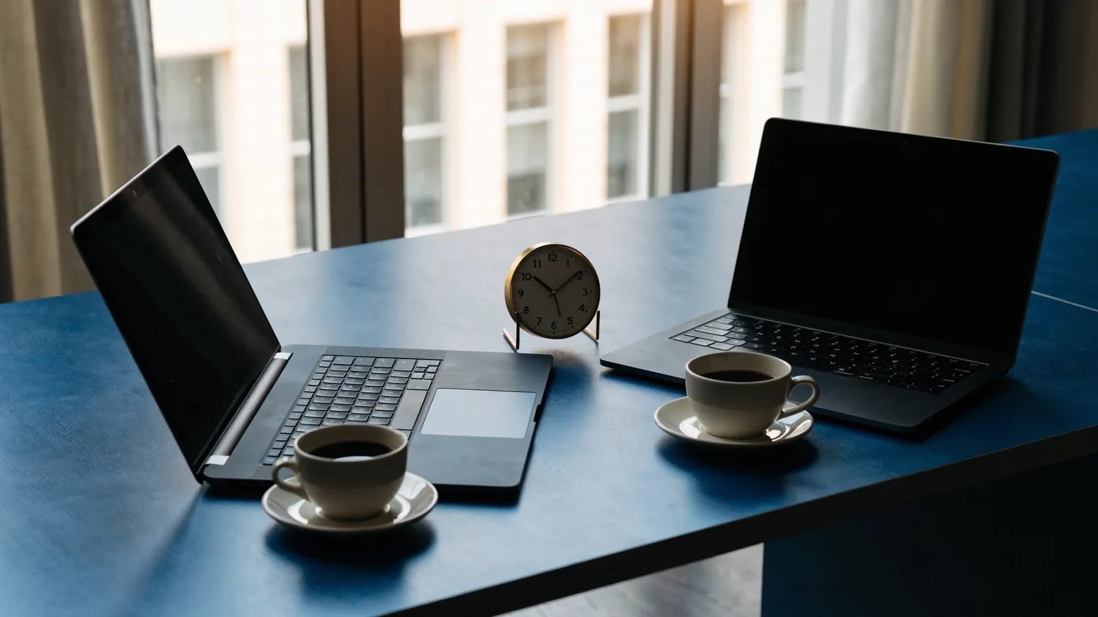 Two desk clocks set to different time zones beside a laptop and a printed contract on a dark wooden desk in window light, no people