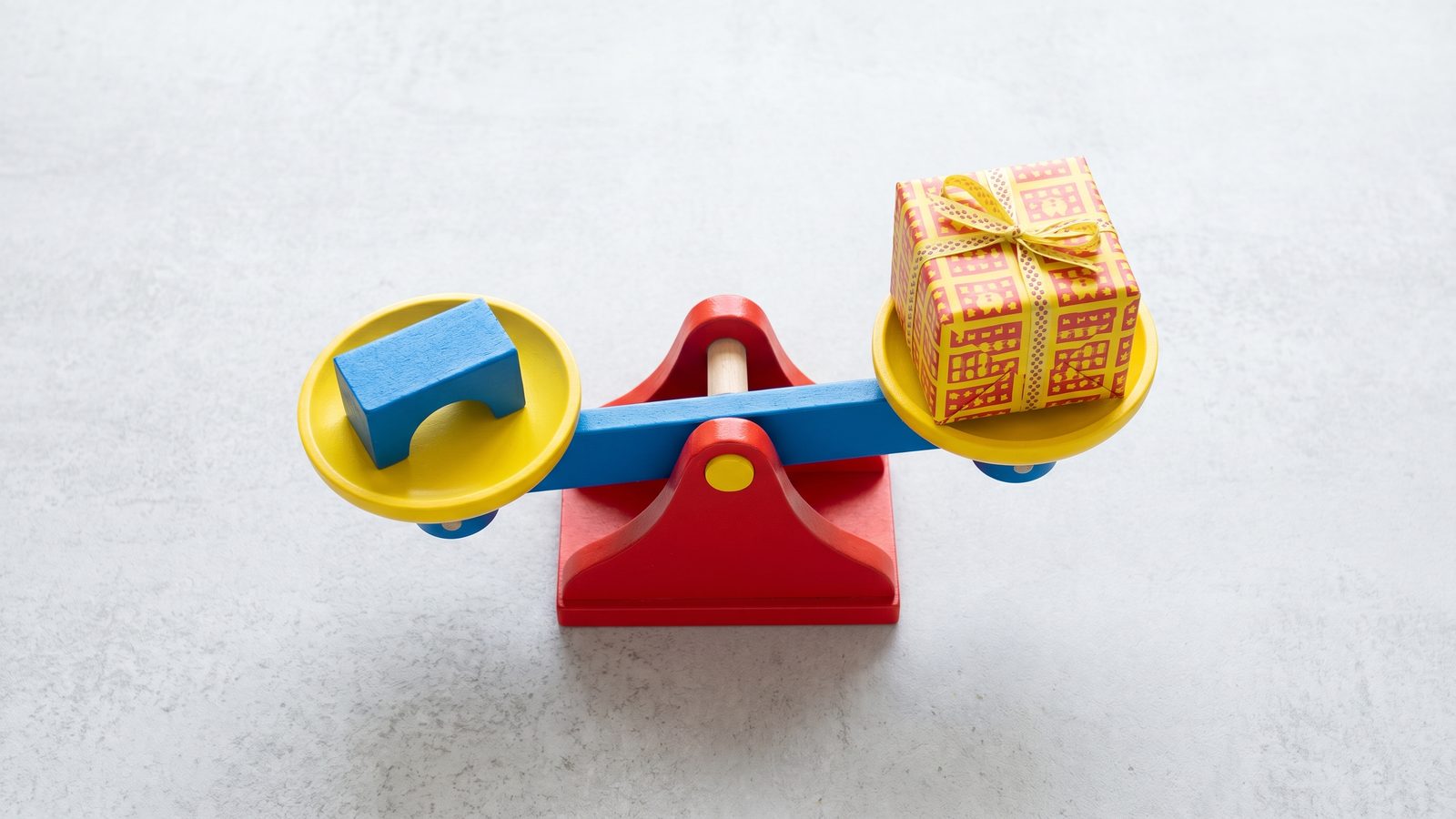 Bright flat lay of a colorful balance scale weighing a building block against a wrapped gift box on a light surface
